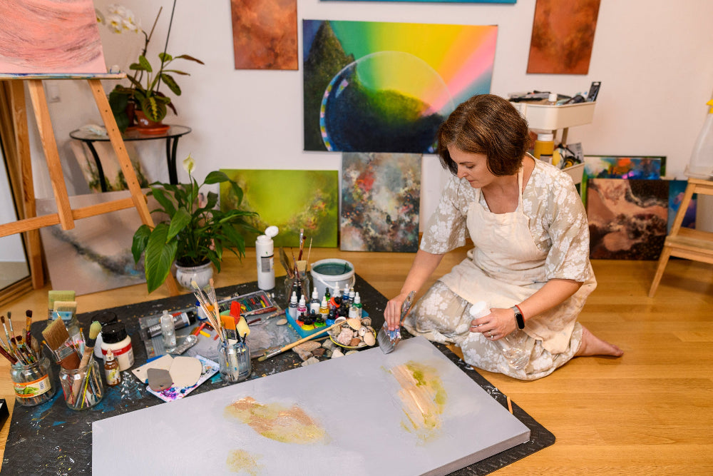 Artist sitting on the floor in an art studio with various paintings and art supplies around.